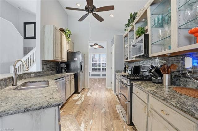 17801 Modena Rd, Miromar Lakes, FL 33913 Kitchen featuring vaulted ceiling, stainless steel appliances, glass insert cabinets, light stone counters, and light wood-style flooring