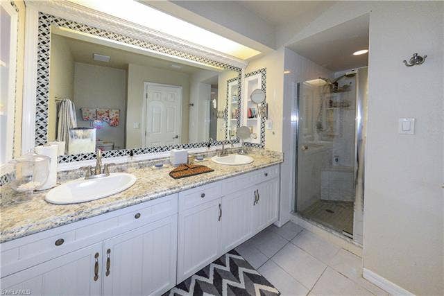 22161 Tallwood Ct, #706, Estero, FL 33928 Bathroom featuring a dual vanity with undermount sinks and a light-toned countertop, a large mirror with decorative tile trim, white cabinetry, a glass-enclosed shower with a built-in bench, and tiled flooring