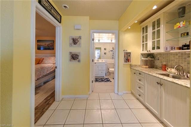 22161 Tallwood Ct, #706, Estero, FL 33928 Hallway featuring white square tile flooring, a built-in wet bar with granite countertops, white cabinetry, a stainless steel sink, and glass shelving