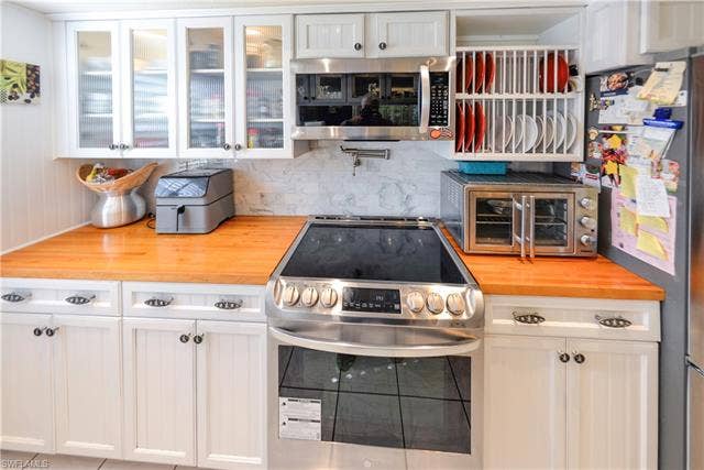 22161 Tallwood Ct, #706, Estero, FL 33928 Modern kitchen featuring a stainless steel range with a flat cooktop, white shaker-style cabinetry, butcher block countertops, a tile backsplash, and upper cabinets with glass-paneled doors