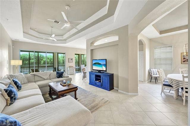 10060 Valiant Ct, #202, Miromar Lakes, FL 33913 Living area featuring large format tile flooring, tray ceiling with dual ceiling fans, expansive sliding glass doors, and arched doorways