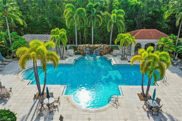 23461 Alamanda Dr, #206, Estero, FL 34135 Expansive outdoor pool area featuring a custom-shaped swimming pool with a rock waterfall feature, surrounding paver patio, a covered cabana with a tile roof, and extensive tropical landscaping