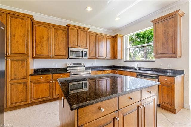 10310 Carolina Willow Dr, Fort Myers, FL 33913 Wood-finish cabinetry with crown molding, dark stone countertops, stainless steel appliances, a kitchen island, and tiled flooring