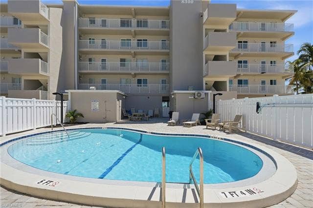 8300 Estero Blvd, #104, Fort Myers Beach, FL 33931 Resort-style saltwater swimming pool featuring lap lanes, curved perimeter design, and stainless steel entry ladder, surrounded by a spacious concrete deck and enclosed by white safety fencing - Virtually Edited Image