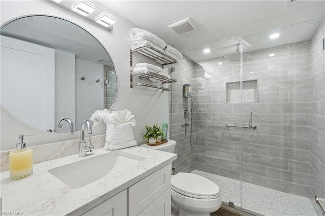 8300 Estero Blvd, #104, Fort Myers Beach, FL 33931 White vanity with integrated sink and chrome faucet, large circular mirror, glass-enclosed shower with tiled walls and built-in niche, and ceiling-mounted showerhead - Virtually Edited Image