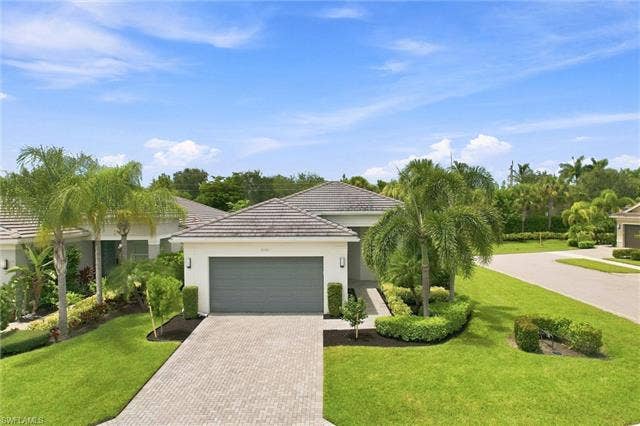 28410 Capraia Dr, Bonita Springs, FL 34135 Tile roof residence featuring an attached garage, paver driveway, manicured lawn, and mature landscaping