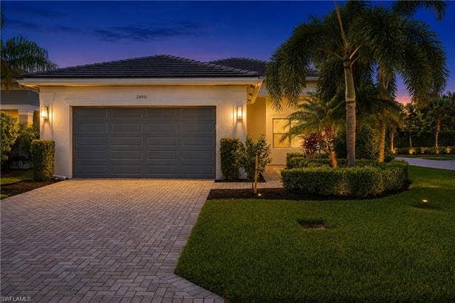 28410 Capraia Dr, Bonita Springs, FL 34135 Paver driveway, exterior wall sconces, integrated garage, manicured shrubbery, and mature palm trees