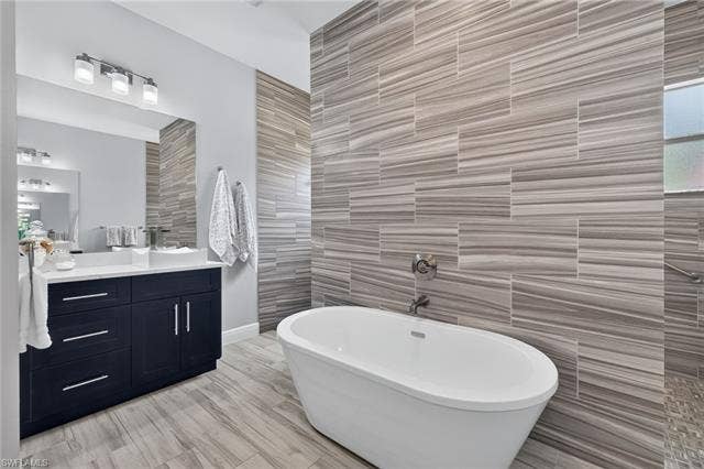 27150 Rue De Paix, Bonita Springs, FL 34135 Freestanding soaking tub, plank tile flooring, dark wood vanities with white quartz countertops, brushed hardware, and tiled accent wall