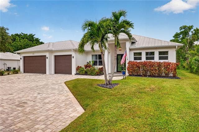 27150 Rue De Paix, Bonita Springs, FL 34135 White stucco exterior featuring a metal roof, three-car garage with brown doors, paver driveway, and mature landscaping