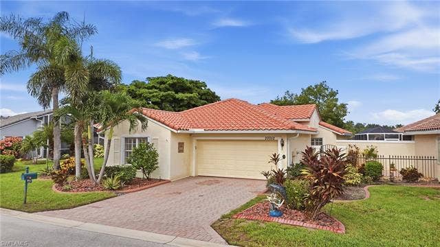20052 Serene Meadow Ln, Estero, FL 33928 Mediterranean-style exterior with a terracotta tile roof, attached garage, paver driveway, mature palm trees, and established landscaping
