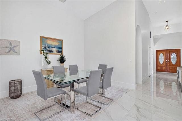 61 Mango St, Fort Myers Beach, FL 33931 Elegant dining area showcasing polished stone flooring, a glass-top table with chrome base and matching upholstered chairs, and abundant natural light from recessed ceiling fixtures - Virtually Edited Image