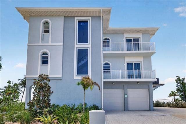 61 Mango St, Fort Myers Beach, FL 33931 Multi-level exterior with light blue siding, multiple balconies with white railings, arched windows, and a paver driveway leading to dual garage doors - Virtually Edited Image
