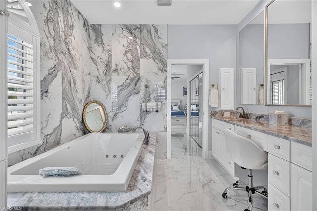 61 Mango St, Fort Myers Beach, FL 33931 Freestanding soaking tub with a tiled surround, marble-finish wall paneling, plantation shutters, dual vanity with undermount sinks, and a makeup counter - Virtually Edited Image