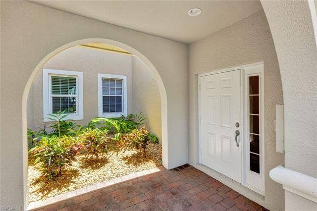10060 Valiant Ct, #202, Miromar Lakes, FL 33913 Arched entryway featuring a white paneled door with sidelight, recessed lighting, brick paver flooring, and an integrated garden area with mature plantings and white-framed windows