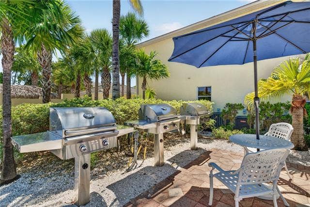 14543 Abaco Lakes Dr, #202, Fort Myers, FL 33908 Community outdoor grilling area featuring multiple stainless steel commercial gas grills, a brick paver patio, and shaded umbrella seating surrounded by tropical palms.
