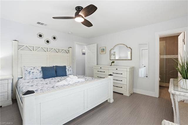 240 Fairweather Ln, Fort Myers Beach, FL 33931 Bedroom featuring light gray walls, wood-finish flooring, a ceiling fan with integrated lighting, and a doorway opening to a shower with tiled walls and a glass enclosure - Virtually Edited Image