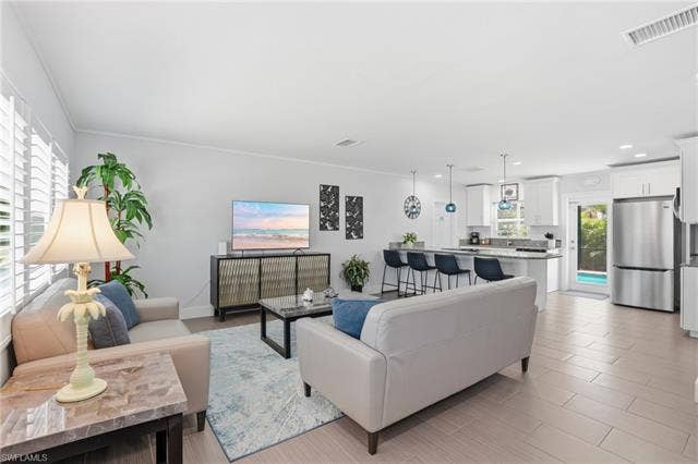 240 Fairweather Ln, Fort Myers Beach, FL 33931 Open-concept living area featuring a kitchen with white cabinetry and stainless steel appliances, a breakfast bar with pendant lighting, and light-toned tile flooring - Virtually Edited Image