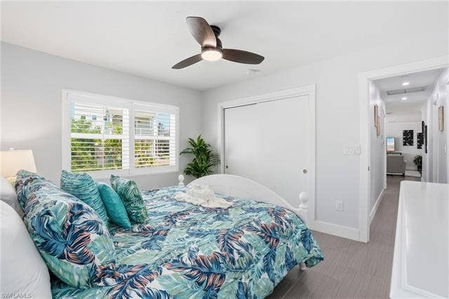 240 Fairweather Ln, Fort Myers Beach, FL 33931 Bedroom featuring a ceiling fan with integrated lighting, a window with louvered shutters, light gray wall paint, and wood-finish flooring - Virtually Edited Image