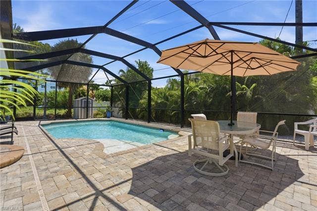 240 Fairweather Ln, Fort Myers Beach, FL 33931 Screened-in pool area featuring a rectangular pool with integrated steps, a paver patio, and mature tropical landscaping beyond the enclosure - Virtually Edited Image