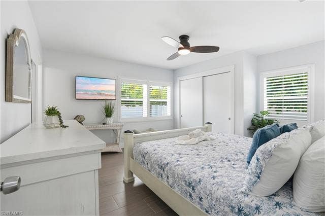 240 Fairweather Ln, Fort Myers Beach, FL 33931 Interior room featuring wood-finish flooring, two windows with plantation shutters, a ceiling fan with integrated lighting, and a closet with sliding doors - Virtually Edited Image