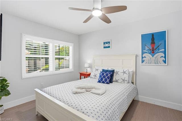 240 Fairweather Ln, Fort Myers Beach, FL 33931 Bedroom featuring light wood-finish flooring, white walls, and a window with plantation shutters - Virtually Edited Image