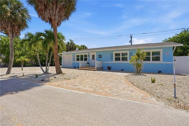 240 Fairweather Ln, Fort Myers Beach, FL 33931 Light blue exterior with white trim, featuring a paver driveway, white privacy fence, and mature palm trees - Virtually Edited Image