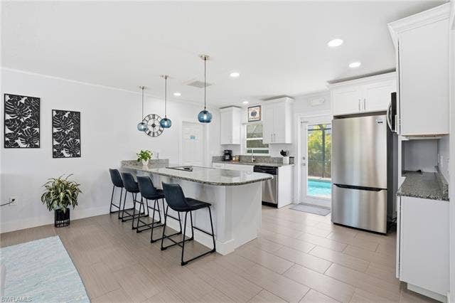 240 Fairweather Ln, Fort Myers Beach, FL 33931 Kitchen featuring an expansive island with seating, white cabinetry, integrated stainless steel appliances, granite countertops, and direct access to an outdoor pool area - Virtually Edited Image