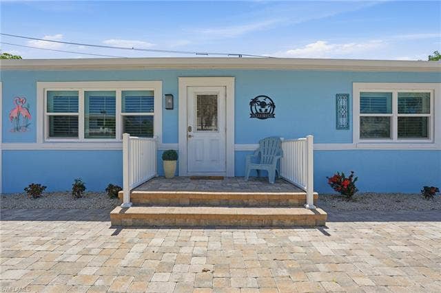 240 Fairweather Ln, Fort Myers Beach, FL 33931 Light blue exterior with white trim, featuring a paver driveway, matching paver porch steps, white railing, and a window with blinds - Virtually Edited Image