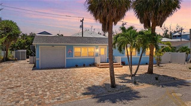 240 Fairweather Ln, Fort Myers Beach, FL 33931 Light blue exterior with white trim featuring a single garage, multiple windows, a front entry with steps, and a paver driveway - Virtually Edited Image