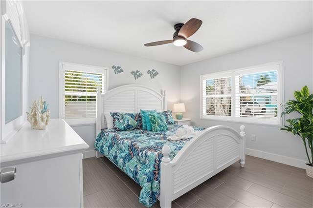 240 Fairweather Ln, Fort Myers Beach, FL 33931 Bright bedroom featuring two windows with white louvered shutters, a white paneled bed frame, a coordinating white dresser with a mirror, a ceiling fan with integrated lighting, and wood-finish flooring - Virtually Edited Image