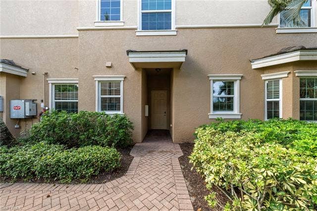 3142 Aviamar Cir, #2104, Naples, FL 34114 Welcoming entry courtyard featuring herringbone paver hardscaping, lush mature landscaping with trimmed hedges, and a neutral stucco facade with multi-pane windows and a covered entryway