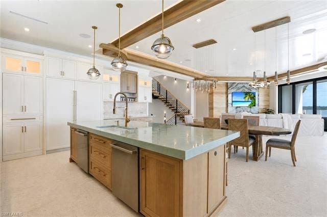 280 Estrellita Dr, Fort Myers Beach, FL 33931 Open-concept kitchen and dining area featuring a substantial island with wood cabinetry and waterfall-edge countertop, stainless steel appliances, white perimeter cabinetry with brass hardware, exposed beam ceiling detail, pendant lighting, and expansive windows with natural light throughout the space - Virtually Edited Image