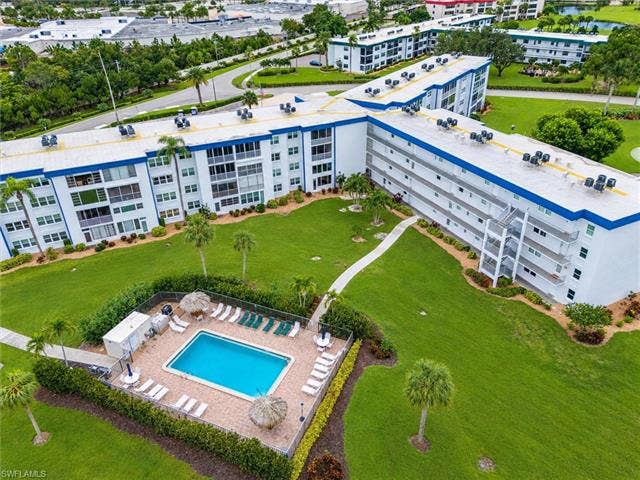 6220 Augusta Dr, #117, Fort Myers, FL 33907 Multi-story residential building featuring white and blue exterior, manicured green lawns, paved walkways, and a fenced swimming pool area with lounge seating
