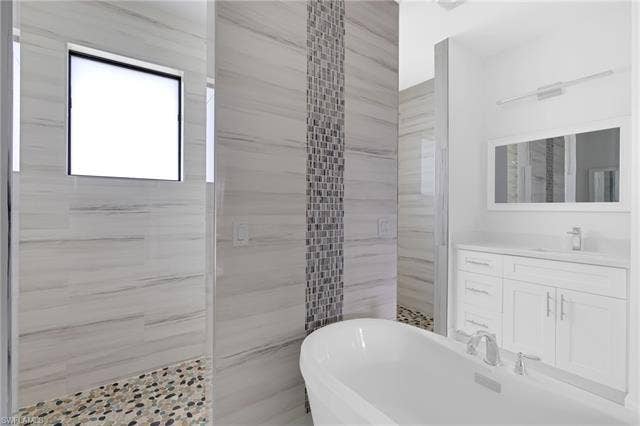 113 38 Pl, Cape Coral, FL 33993 Freestanding soaking tub, walk-in shower with pebble tile flooring, white vanity with integrated sink, rectangular framed mirror, and a mosaic tile accent wall