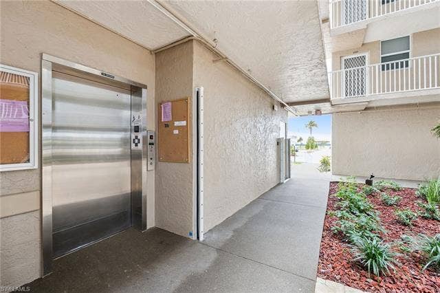 5500 Bonita Beach Rd, #308, Bonita Springs, FL 34134 Stainless steel elevator with call buttons, textured exterior walls, concrete walkway, and landscaped garden beds with red mulch