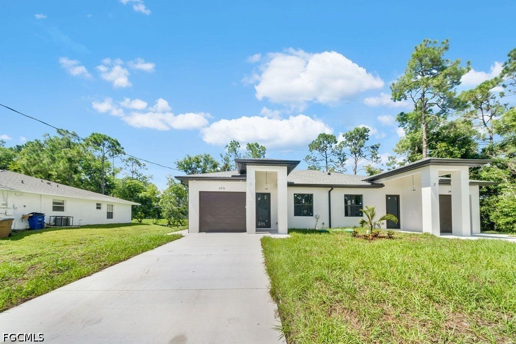 2416/2418 Joe Avenue S, Lehigh Acres, FL 33973 Contemporary exterior featuring a light-colored facade, dark-framed windows, an attached garage, a concrete driveway, and a landscaped front yard
