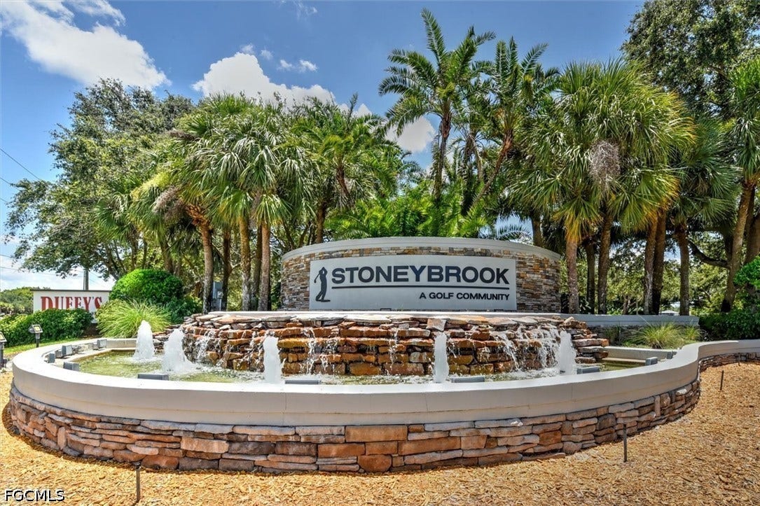 Community entrance featuring a stone-clad sign, layered stone fountain, and mature palm trees