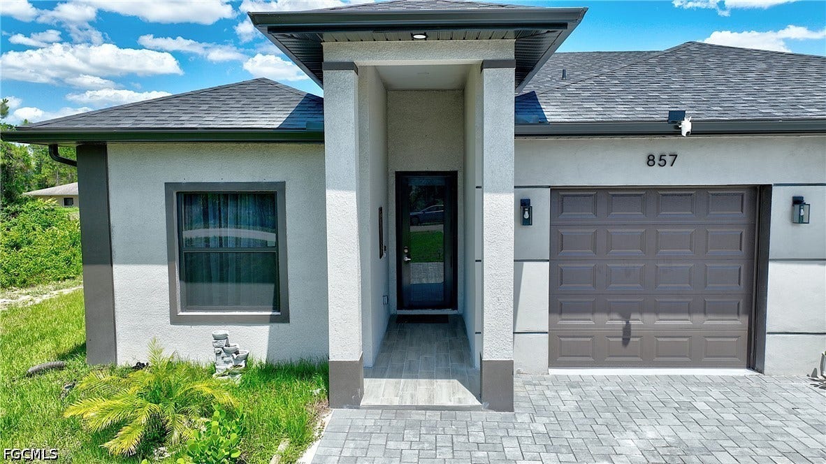 View of exterior entry featuring roof with shingles, stucco siding, a garage, and driveway