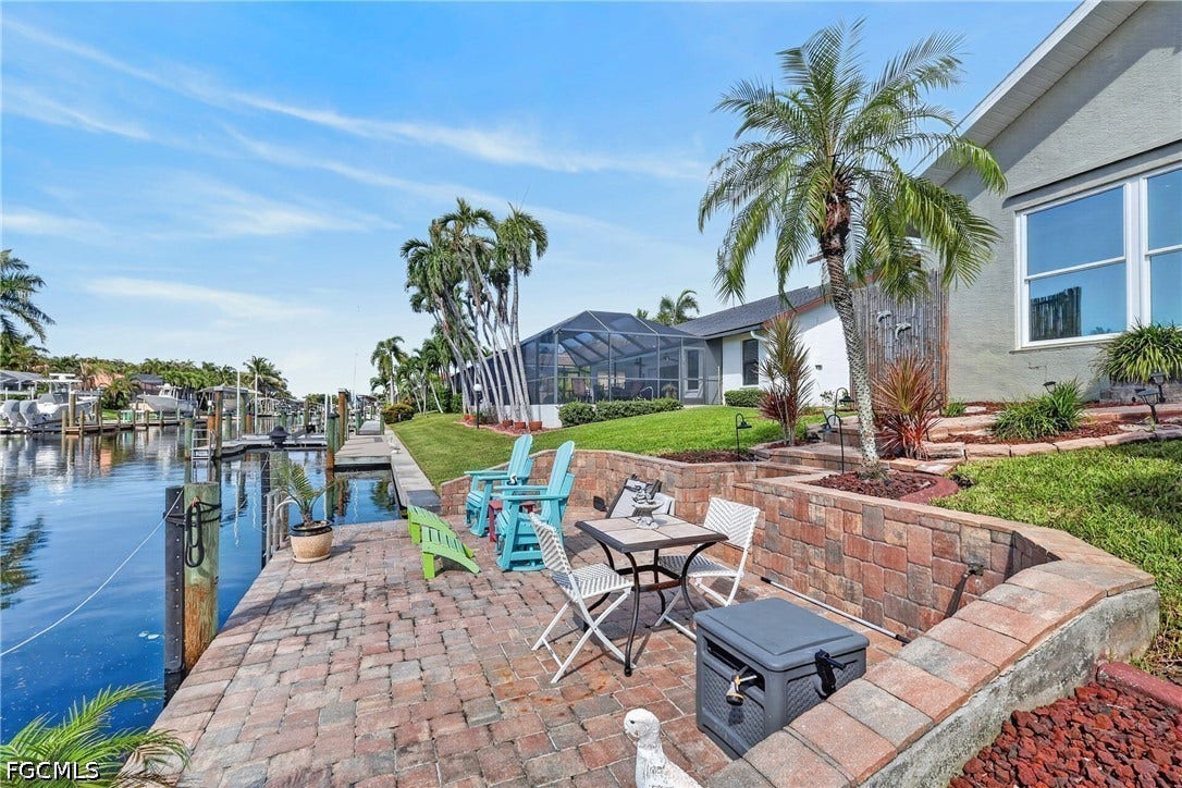 1416 SW 49th Terrace, Cape Coral, FL 33914 Waterfront property featuring a brick paver patio, a tiered landscaping design, mature palm trees, and a screened enclosure