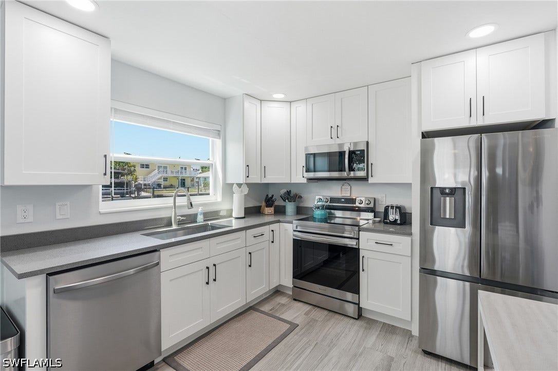 Modern kitchen featuring white shaker cabinetry, stainless steel appliances, gray countertops, a stainless steel sink with a gooseneck faucet, and light wood-finish flooring - Virtually Edited Image