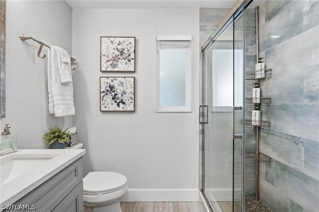 Contemporary bathroom featuring a glass-enclosed shower with tile surround, a single vanity with a solid surface countertop, wood-finish flooring, and a window providing natural light - Virtually Edited Image