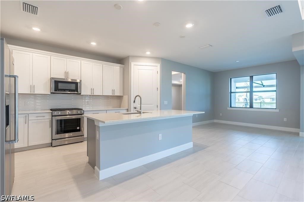 Open-concept kitchen featuring white shaker cabinetry, stainless steel appliances, a center island with a sink, and light-toned tile flooring