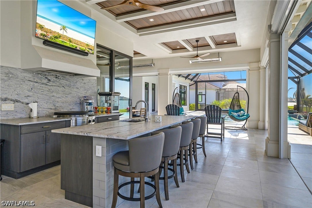 Outdoor kitchen with leathered quartz countertops, built-in cabinetry, EVO affinity flat top grill, beverage cooler, ice maker, stainless steel outdoor sink, and TV with sound bar.