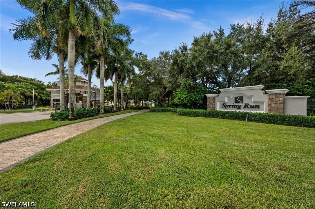 Gated community entrance with stone-accented signage, mature landscaping, and a paver pathway