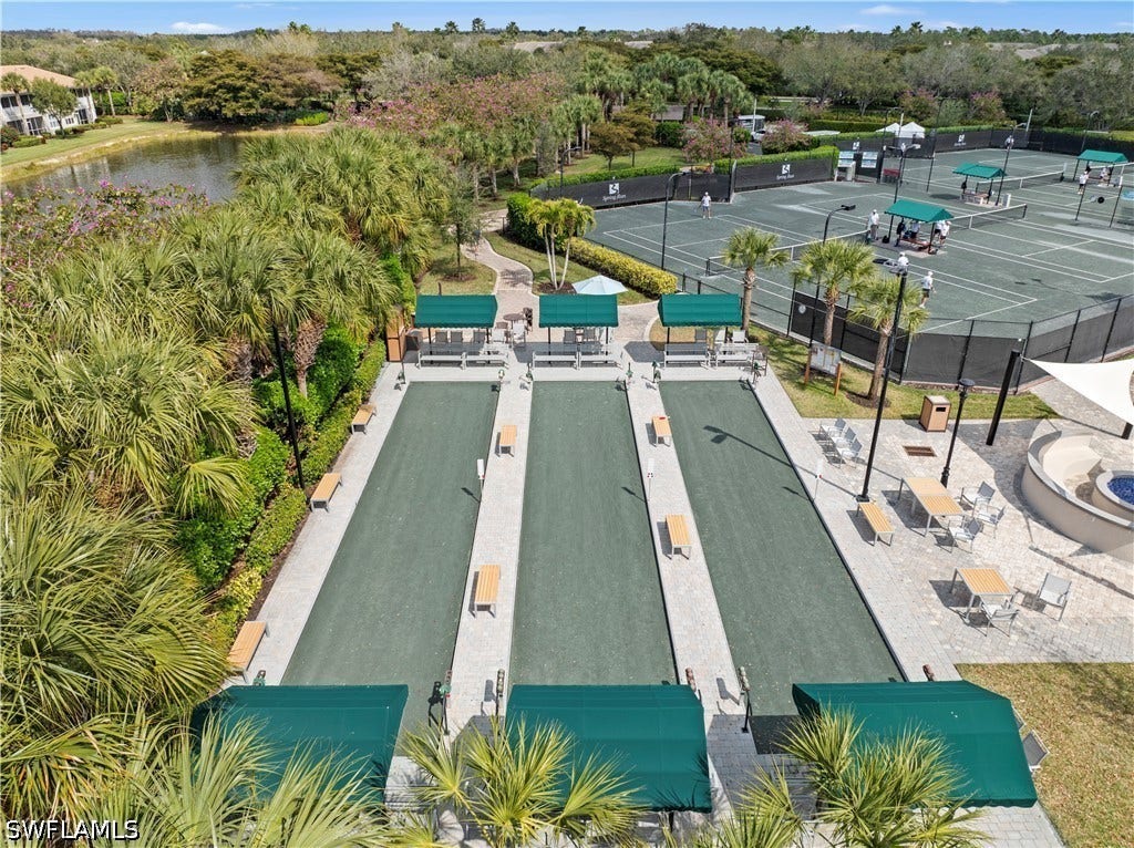 Multiple bocce ball courts with shaded seating, tennis courts with a dark perimeter fence, a fire pit area, and a tranquil pond bordered by lush tropical landscaping