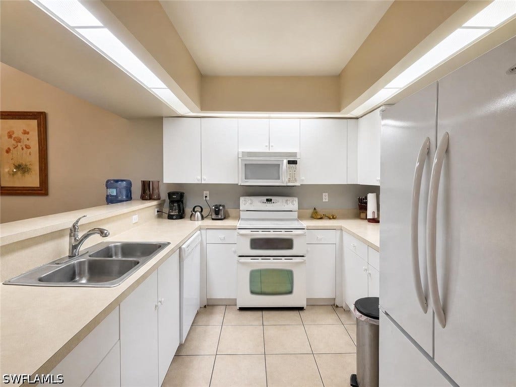 U-shaped kitchen featuring white cabinetry, light-toned countertops, a double basin stainless steel sink, and white appliances