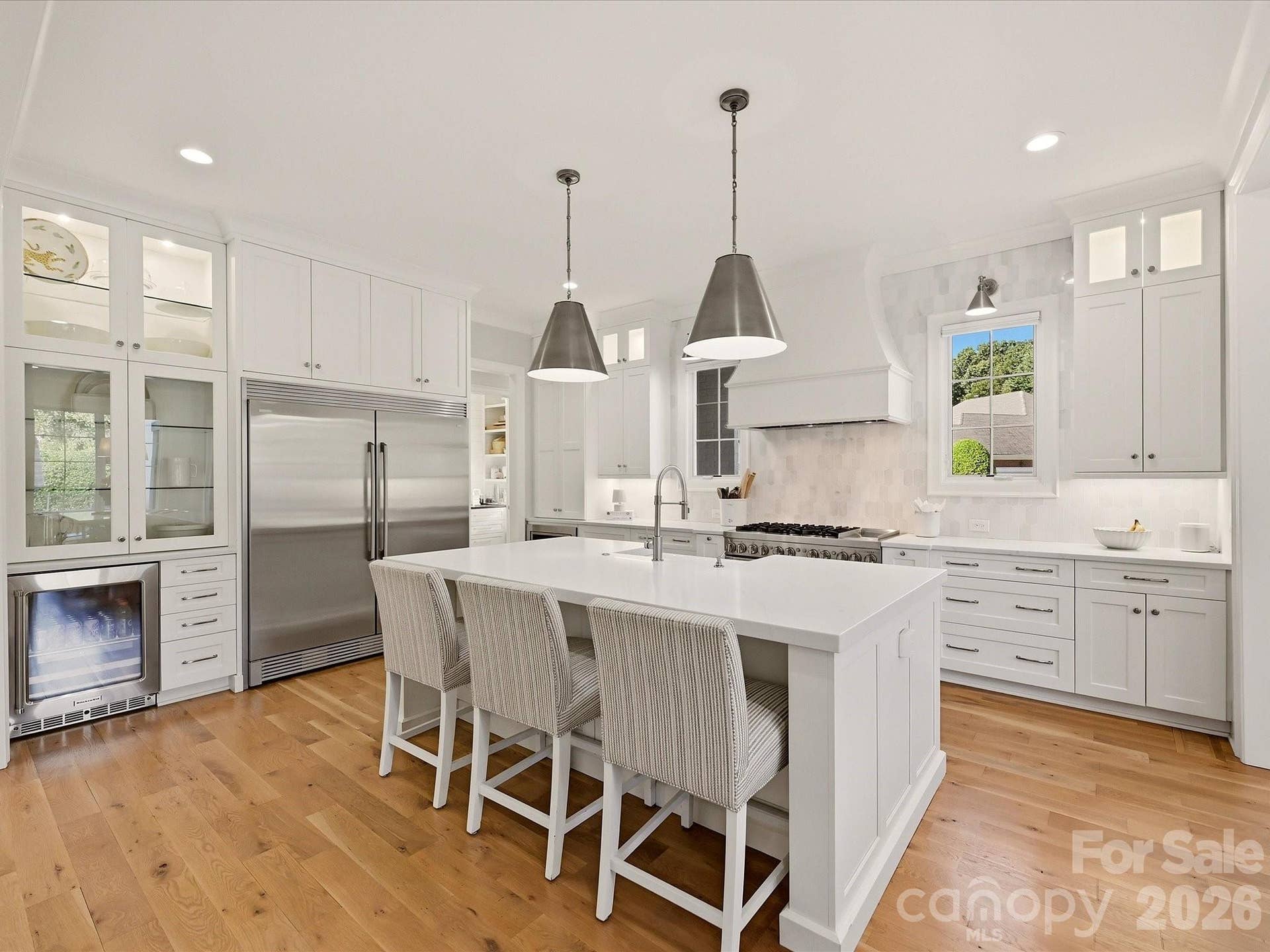 Bright designer kitchen features an oversized island with seating, custom cabinetry, stainless steel appliances, a statement range hood, beverage refrigerator, and abundant workspace for everyday living and entertaining.