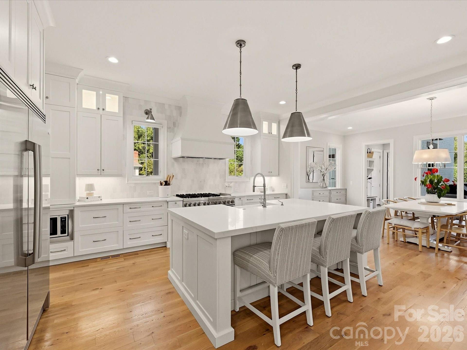 Bright designer kitchen features an oversized island with seating, custom cabinetry, stainless steel appliances, a statement range hood, beverage refrigerator, and abundant workspace for everyday living and entertaining.