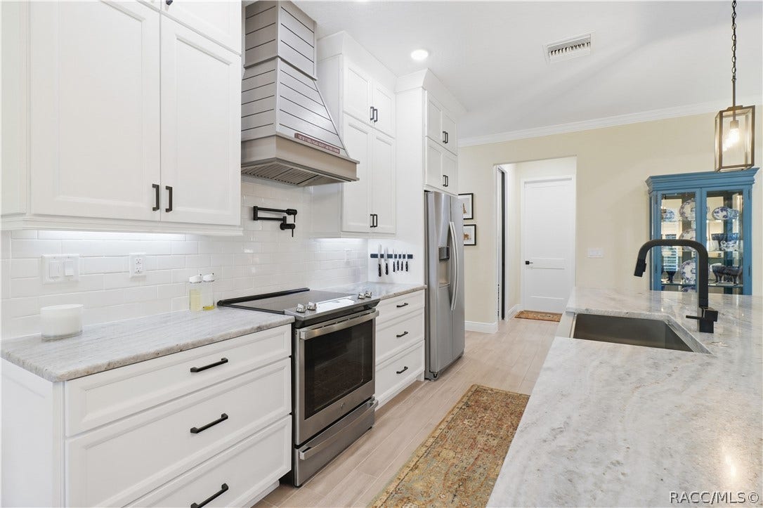 4124 W Pine Ridge Boulevard, Beverly Hills, FL 34465 DESIGNER KITCHEN Tall white cabinetry, black hardware, under-cabinet lighting, subway-tile backsplash, stainless appliances, custom-style hood & pot filler combine style w/ everyday function.