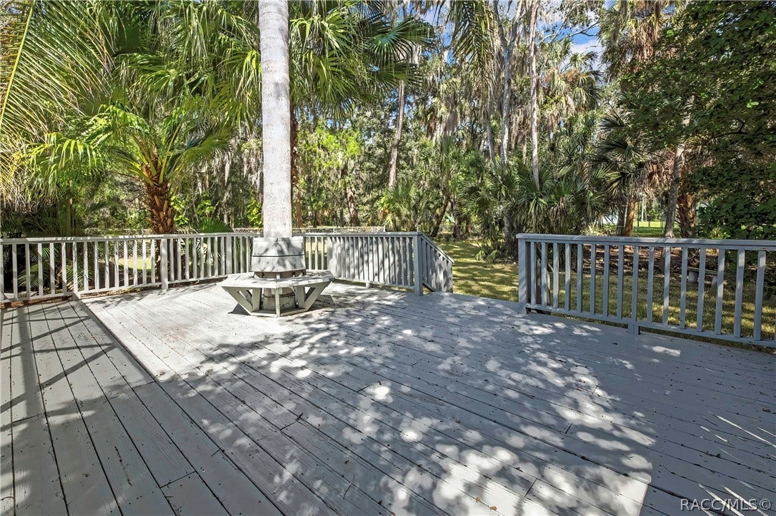 4737 S Sawmill Way, Homosassa, FL 34448 Wood deck: grey wood deck planks, a built-in bench around a palm tree trunk in the centre, railing and steps at the left, the screen enclosure at the right, trees, and a neighbouring house in the background.